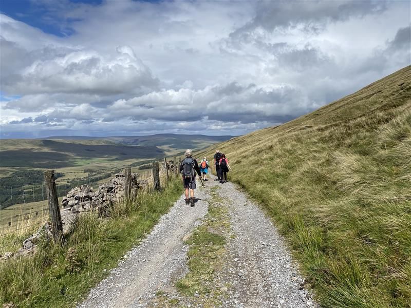 A group of ramblers walk along a gravel footpath along the Yorkshire countryside, on a cloudy day. They walk away from the camera towards the horizon. Clouds roll across the hills and green hills expand either side of the footpath